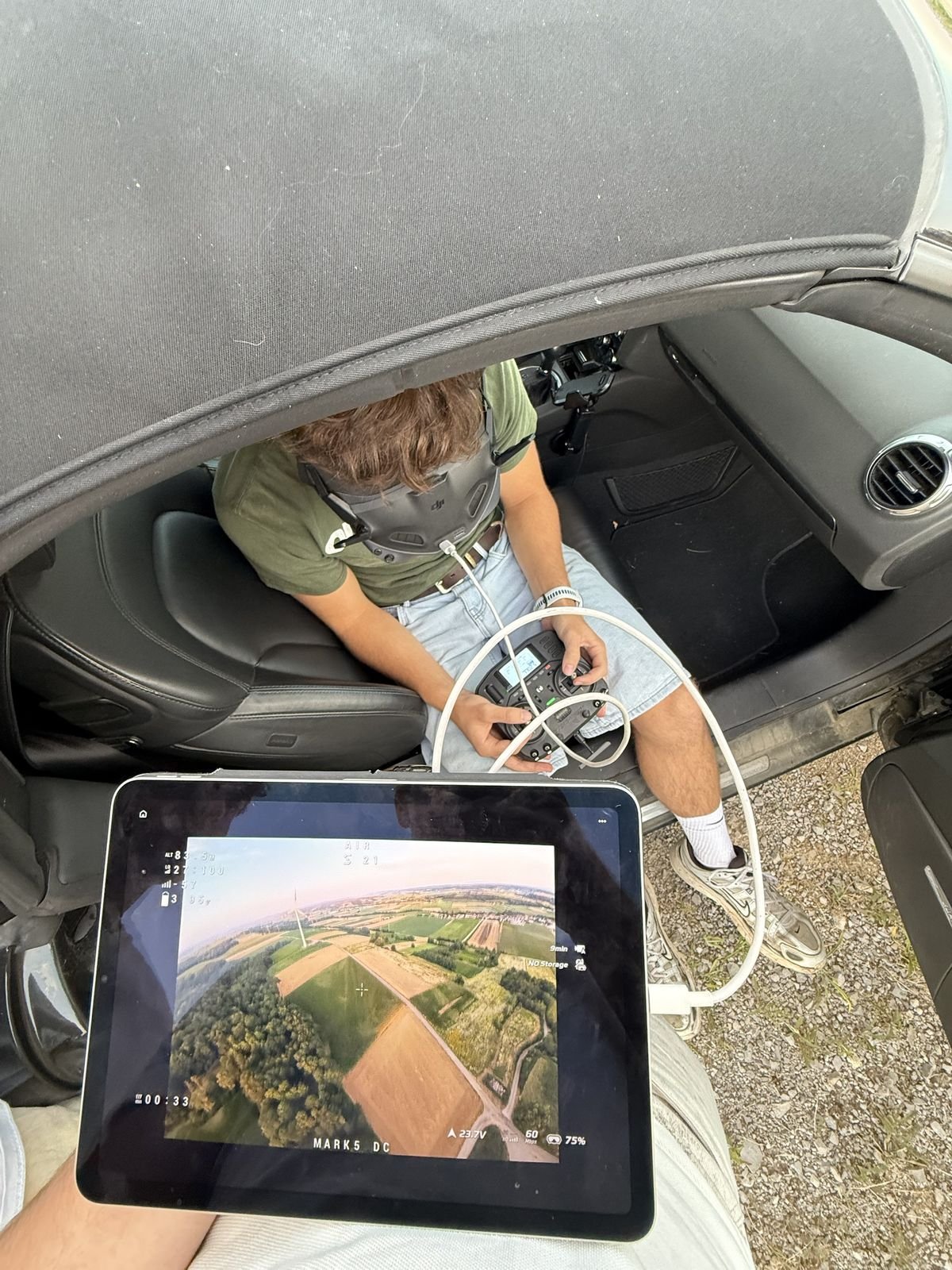Pilot mit FPV-Brille und Fernsteuerung im Auto, auf dem iPad das Live-Bild der Drohne